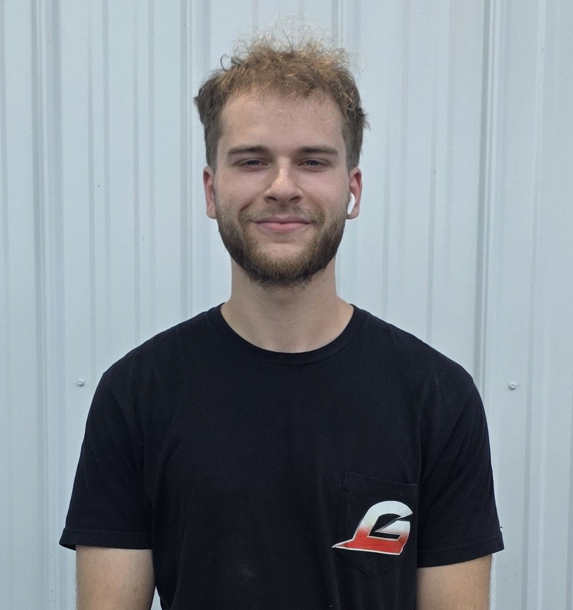 Smiling person with short hair wearing a black T-shirt against a light corrugated wall