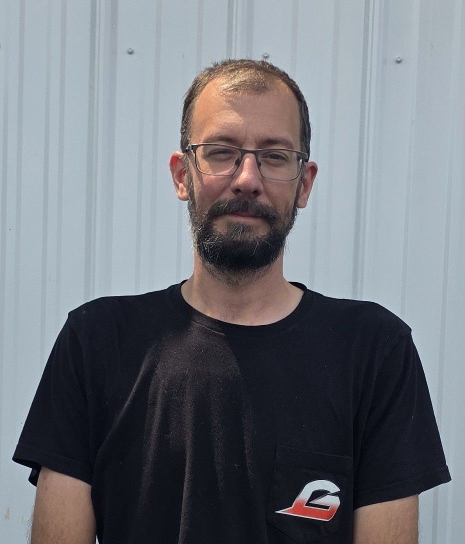 Man standing against a light corrugated wall, wearing a black T-shirt and glasses, with a neutral expression