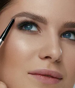 Close-up of a woman applying eyebrow makeup with a brush, showing blue eyes and soft pink lips