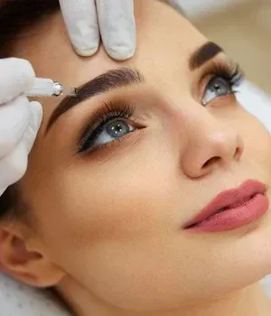 Gloved hands filling a woman's eyebrow with a cosmetic tattoo pen, close-up of her face