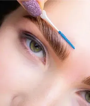 Applying a blue brow gel with a wand to a green eye makeup close-up