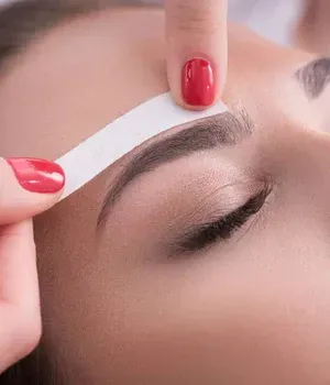 Hands waxing a eyebrow with white strip and red nails on a closed eye.