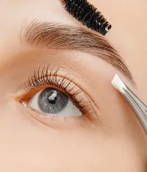 Close-up of an eye while a mascara wand and tweezers groom the eyebrow