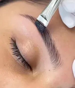 Brush applying dark tint to an eyebrow on a closed eye, close-up beauty treatment
