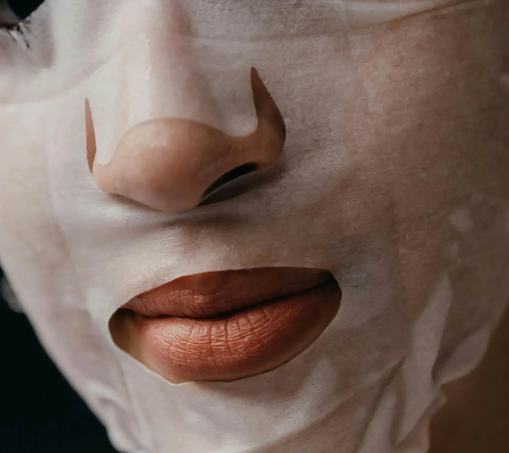Close-up of lips and nose covered by a white facial sheet mask, showing a cutout around the mouth.