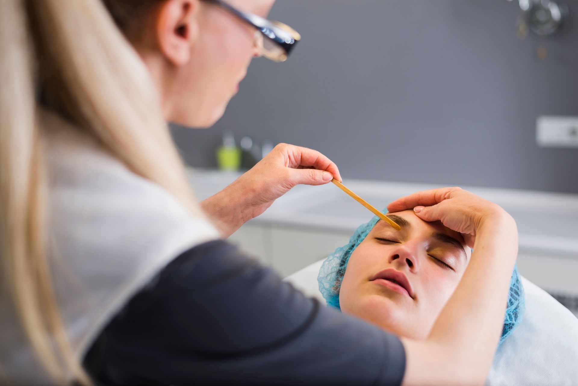 A beautician applies a facial treatment to a client lying on a bed with a blue headband.