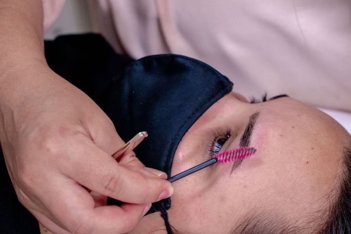 Person receiving eyelash extension while lying down, with tweezers and pink spoolie near the eye