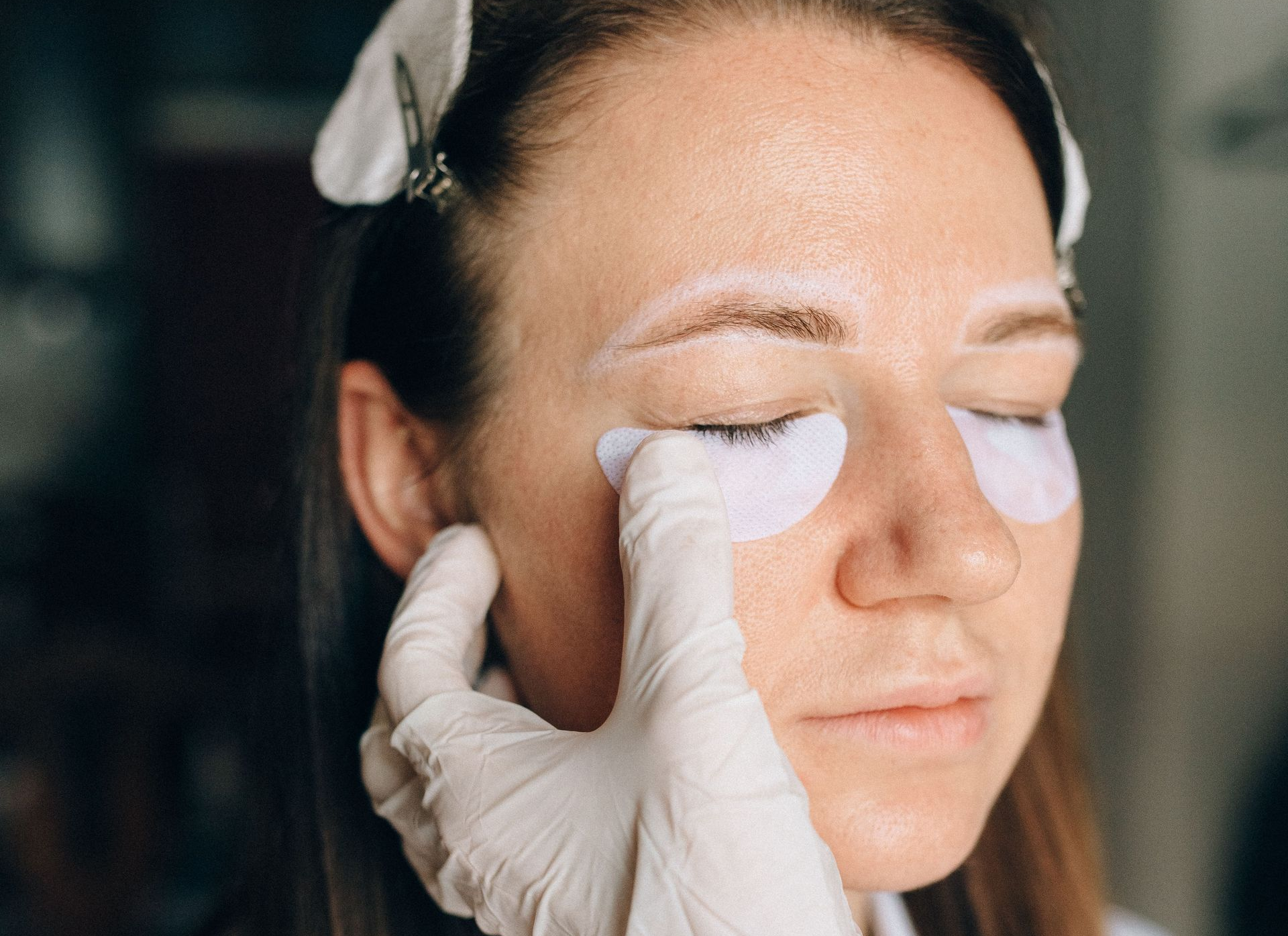 Gloved hand applying a white pad to a woman’s closed eye during a facial treatment