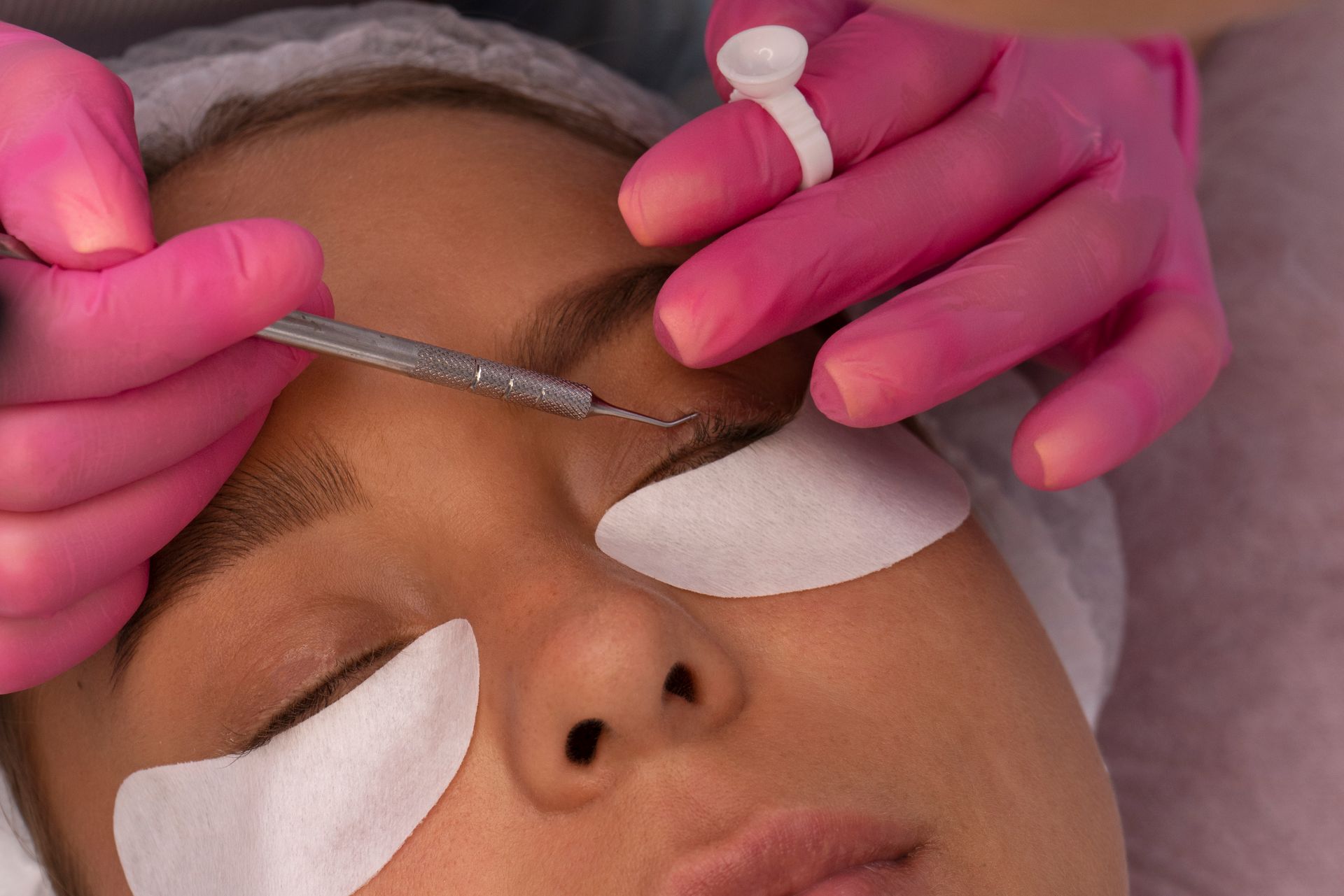 Cosmetic treatment on eyebrow area with hands holding tools and eye pads in place
