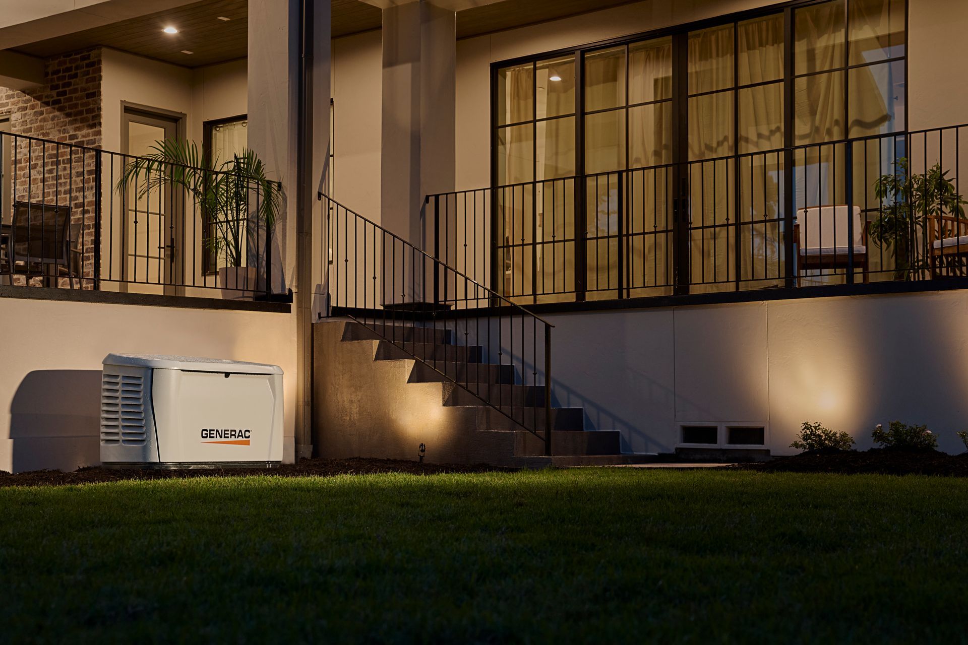 A Generac home standby generator sits on the lawn next to the steps of a modern house illuminated at night.