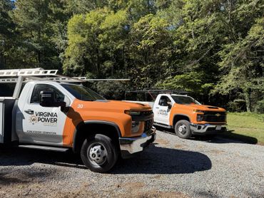 Two orange-and-white Virginia Power utility trucks parked on gravel by trees