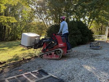 Person on red mini skid steer working on gravel driveway beside trees and a covered utility trailer.