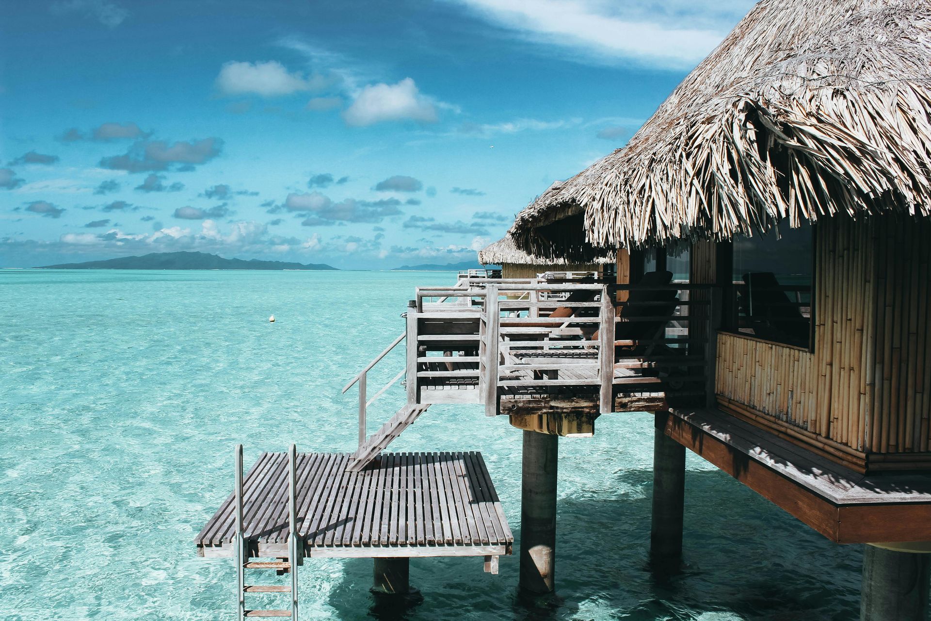 Overwater bungalow with thatched roof and deck, turquoise ocean, clear sky.