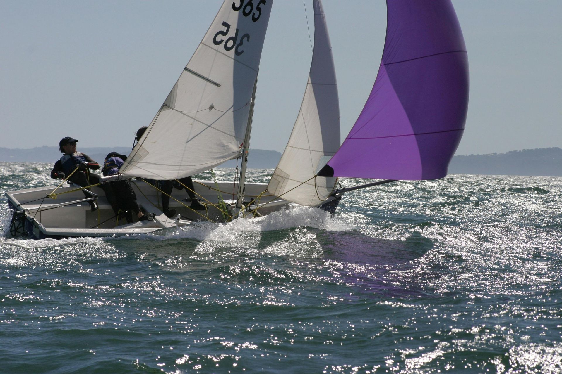 Sailboat racing on choppy water with purple sail raised; two people on board.