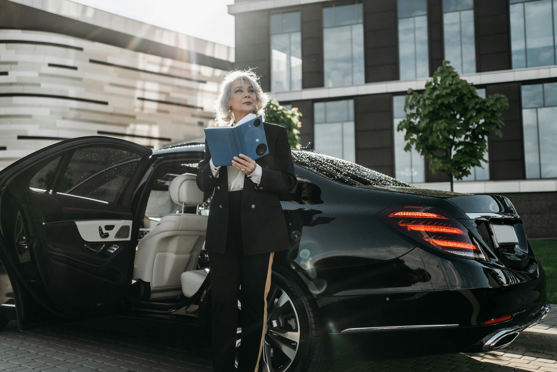 Woman in a suit stands by a black luxury car, holding a notebook, in front of a modern building.