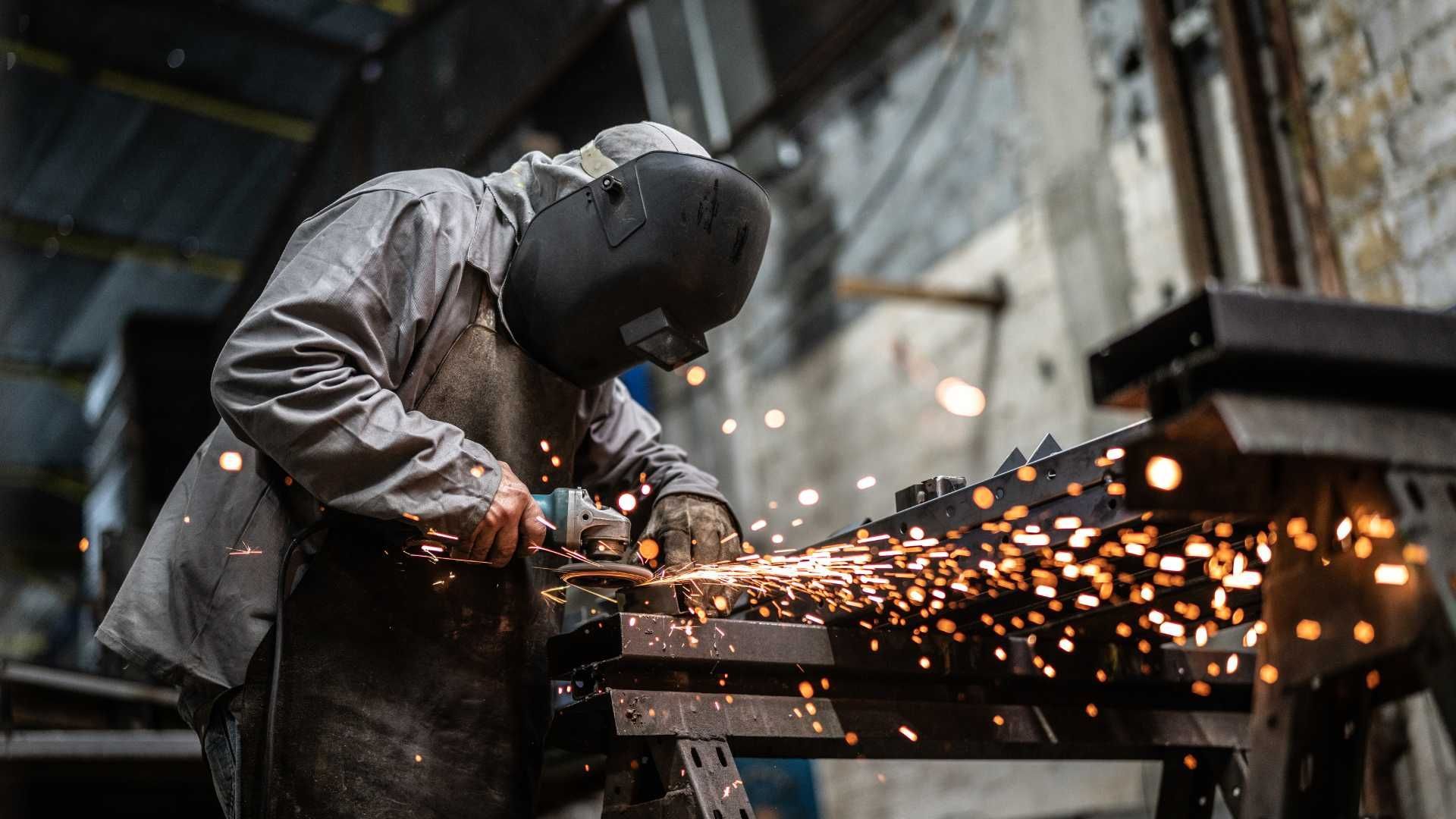 A man wearing a welding mask is grinding a piece of metal in a factory.