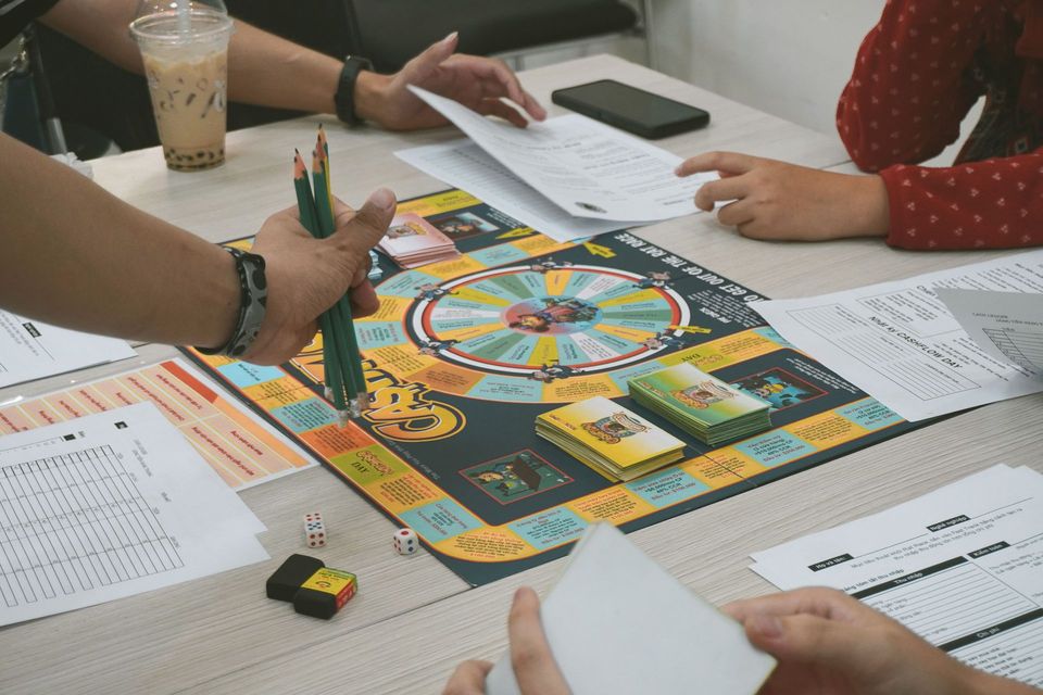 People playing a colorful board game at a table with dice, cards, and papers