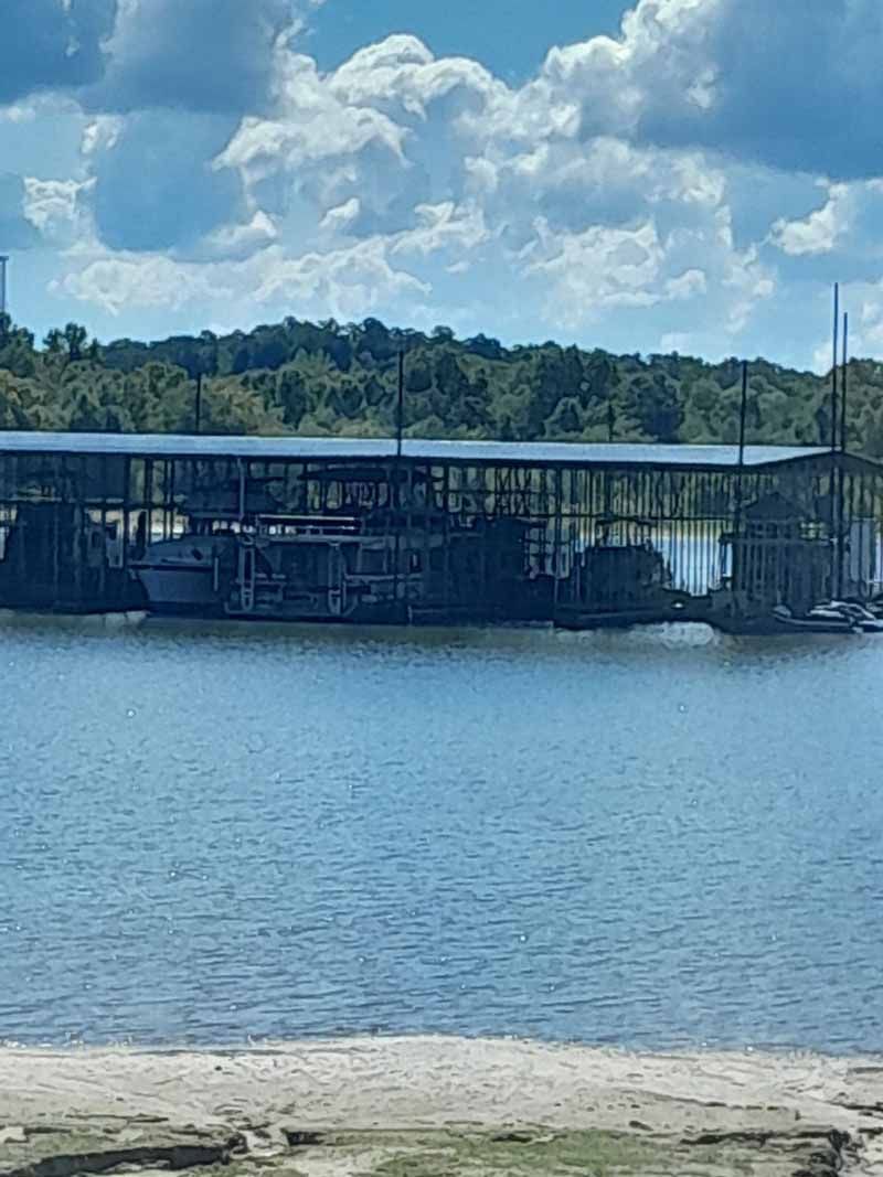 Boats docked under a covered structure on a body of water. Blue sky and trees in the background.