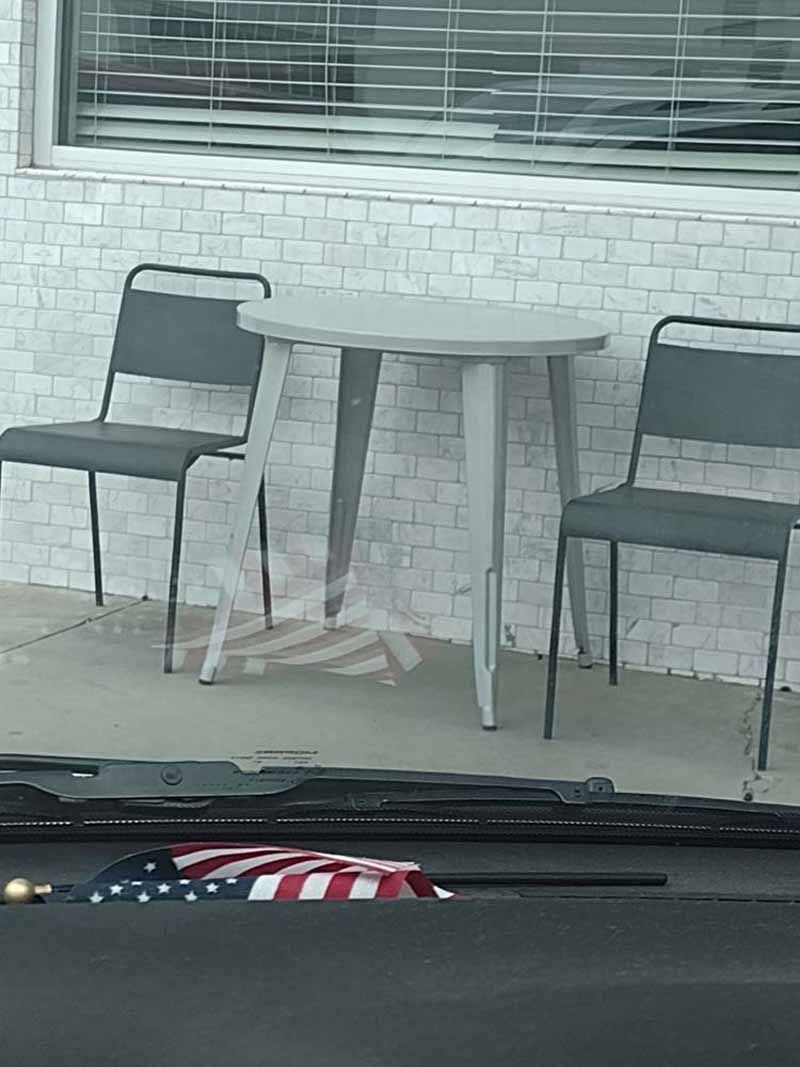Two chairs and a table on a patio, in front of a window with blinds. An American flag is in the foreground.