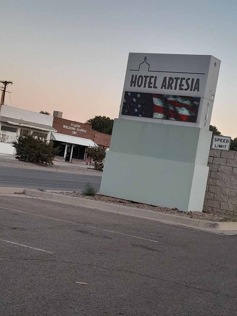 Hotel Artesia sign on a roadside, light blue sky. Building with an American flag displayed on the sign.