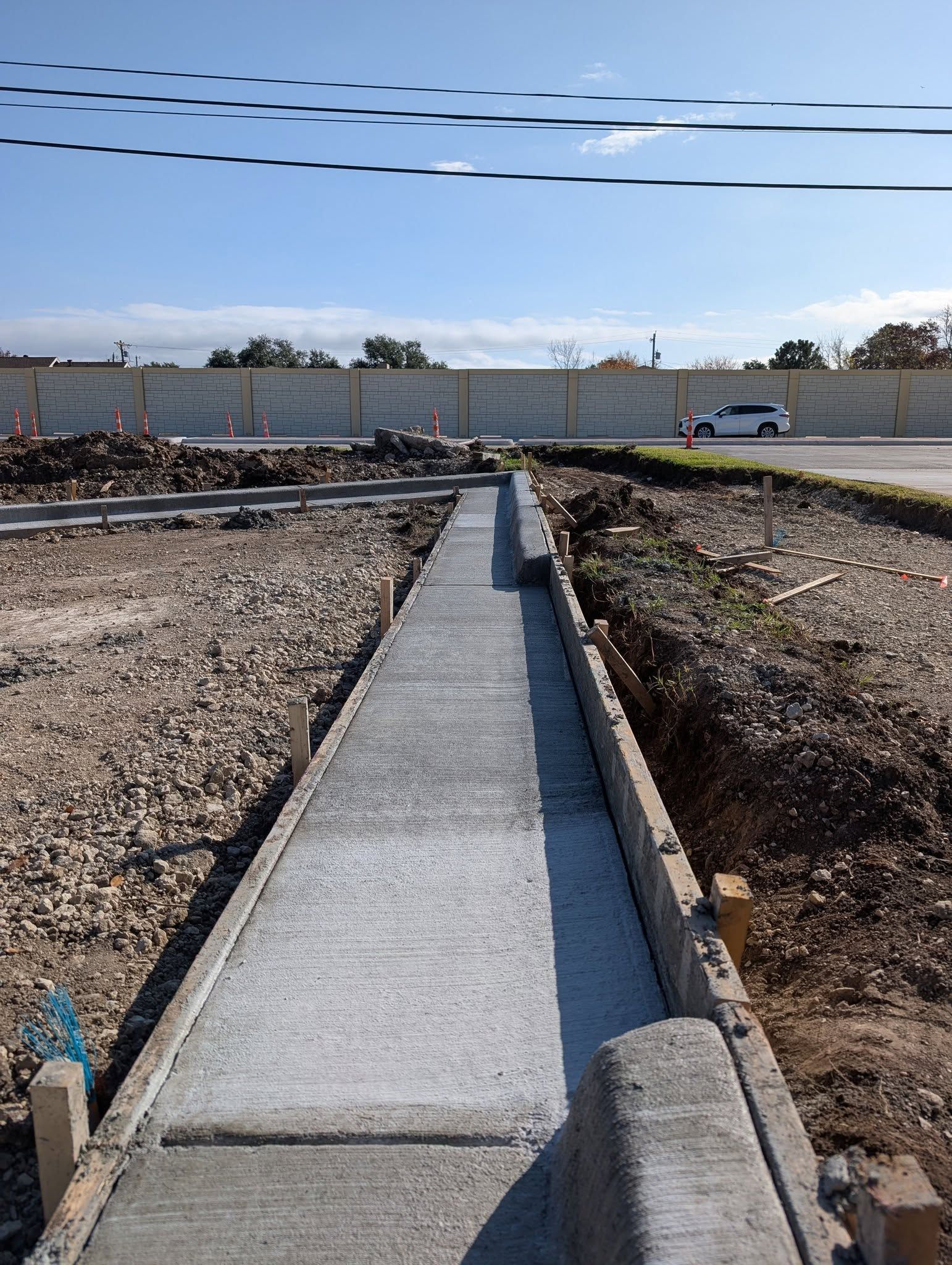 Newly poured concrete sidewalk with wood forms alongside a road, under a blue sky.