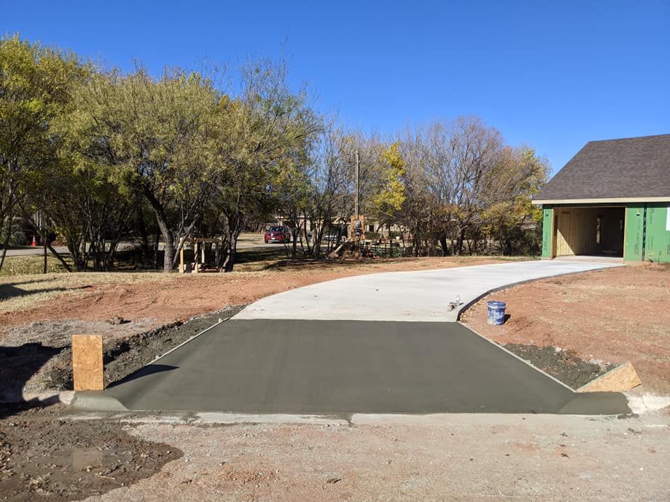 New concrete driveway and entrance under construction next to a house with a garage and a tree-lined yard.