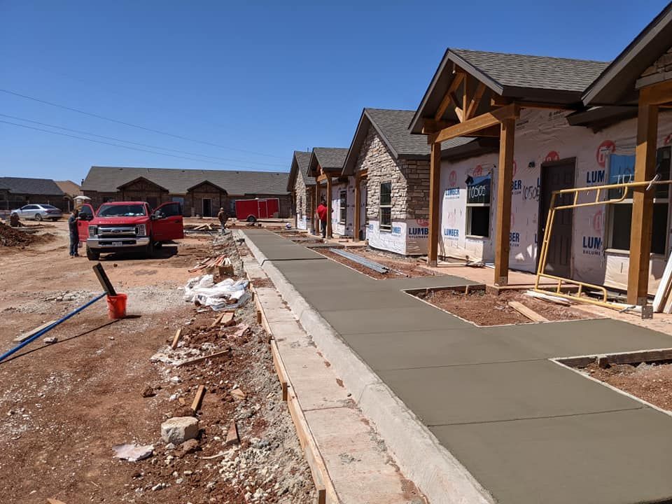 Construction site with new concrete sidewalk being poured in front of multi-unit buildings. Red truck and workers present.