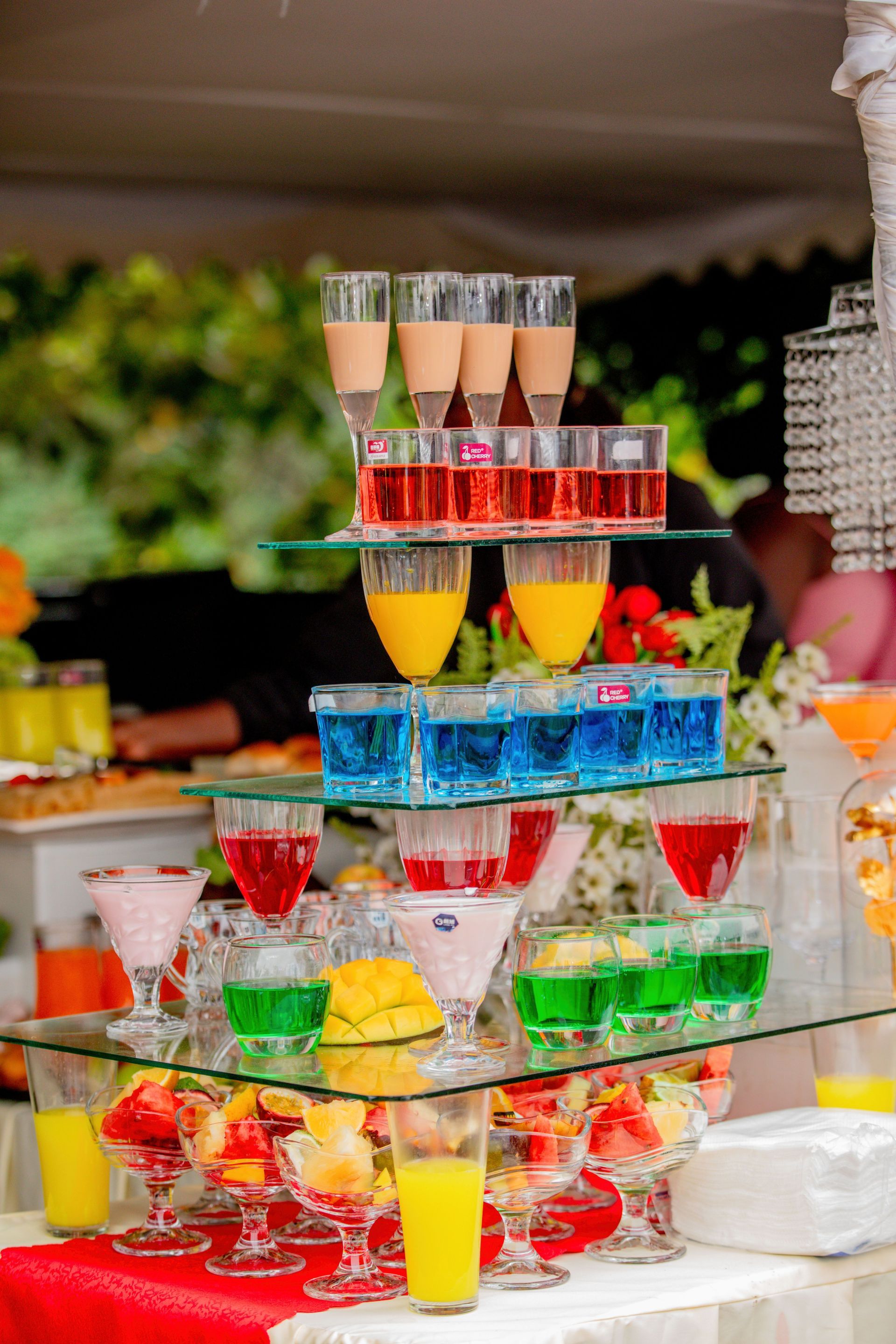 Tiered tray of colorful layered dessert shots in red, yellow, blue, and green at a buffet table