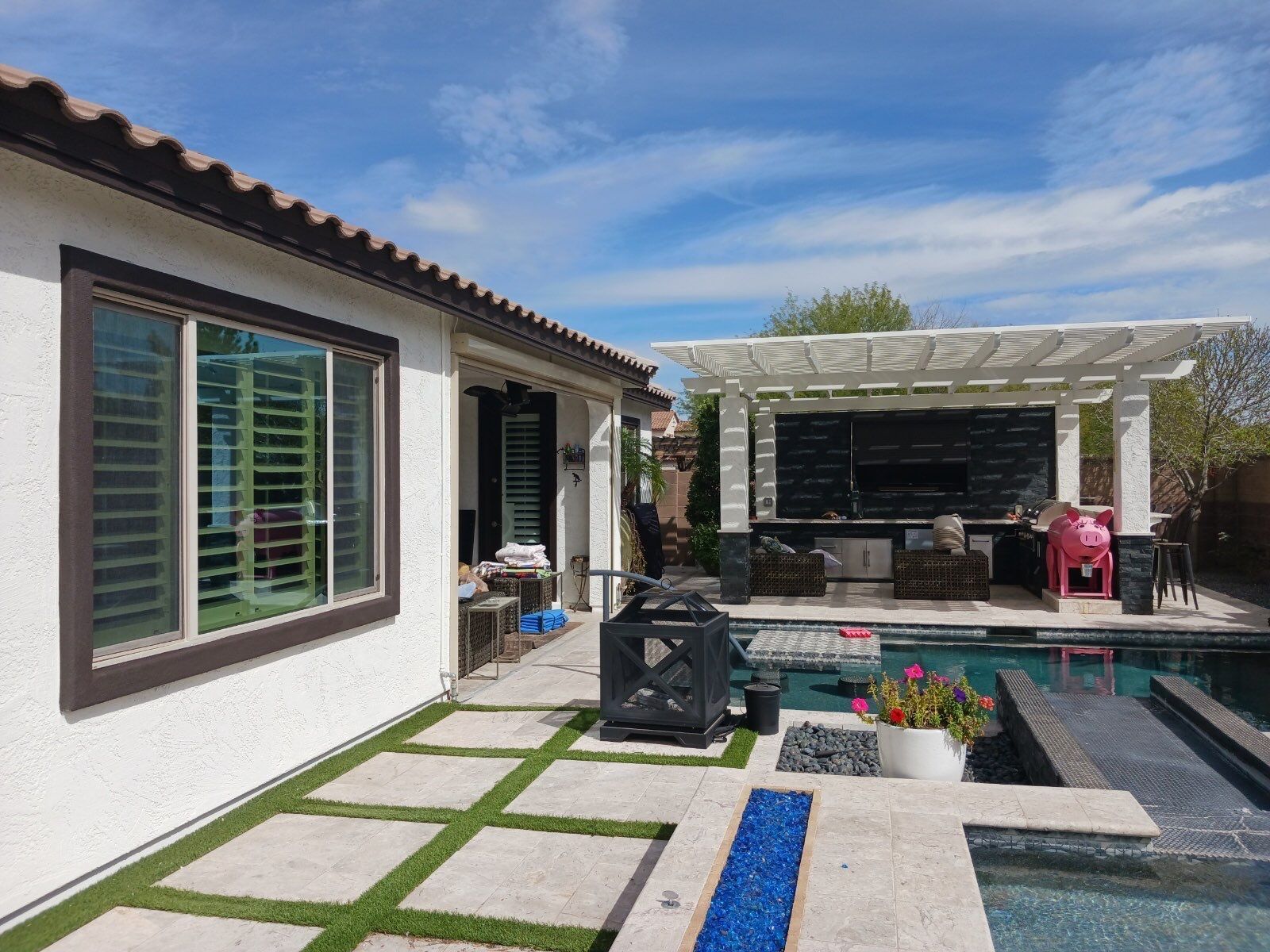 Backyard with pool, outdoor kitchen, patio. White house with brown trim. Blue sky, sunny day.