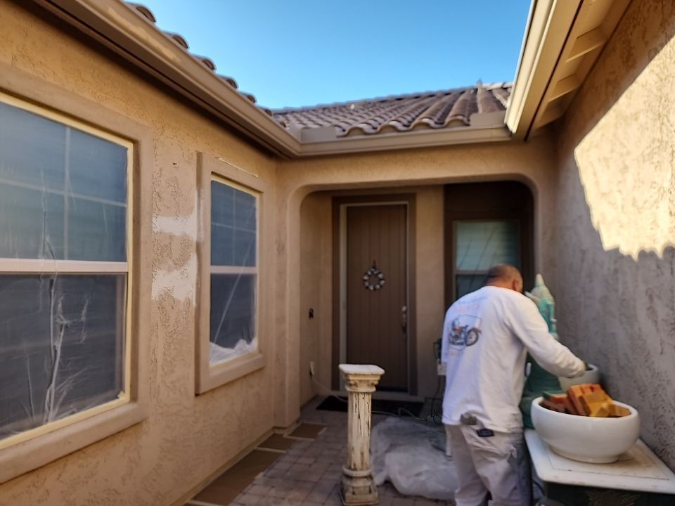 A man paints an exterior stucco wall.  A tan house with brown trim and tile roof.