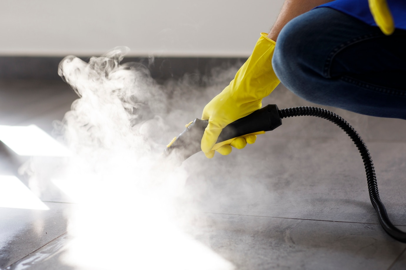 A person wearing yellow gloves is using a steam cleaner on a tiled floor.