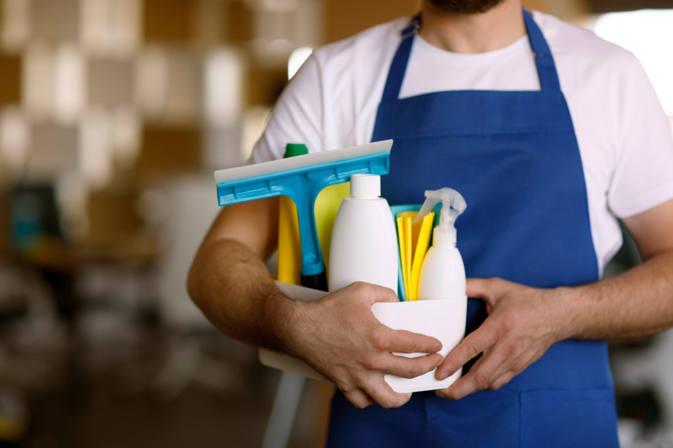 A man in an apron is holding a basket of cleaning supplies.
