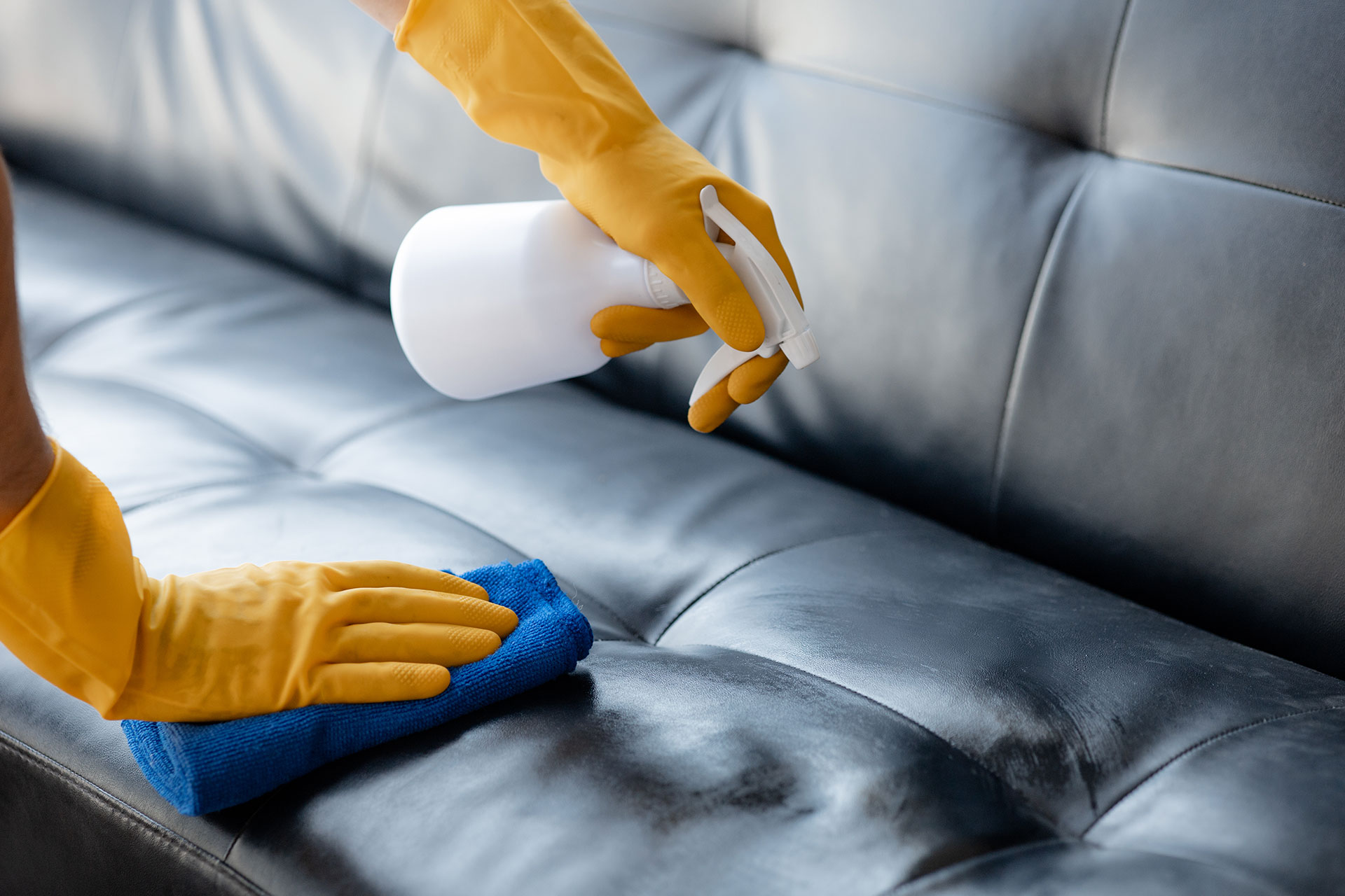 A person wearing yellow gloves is cleaning a leather couch with a blue cloth.