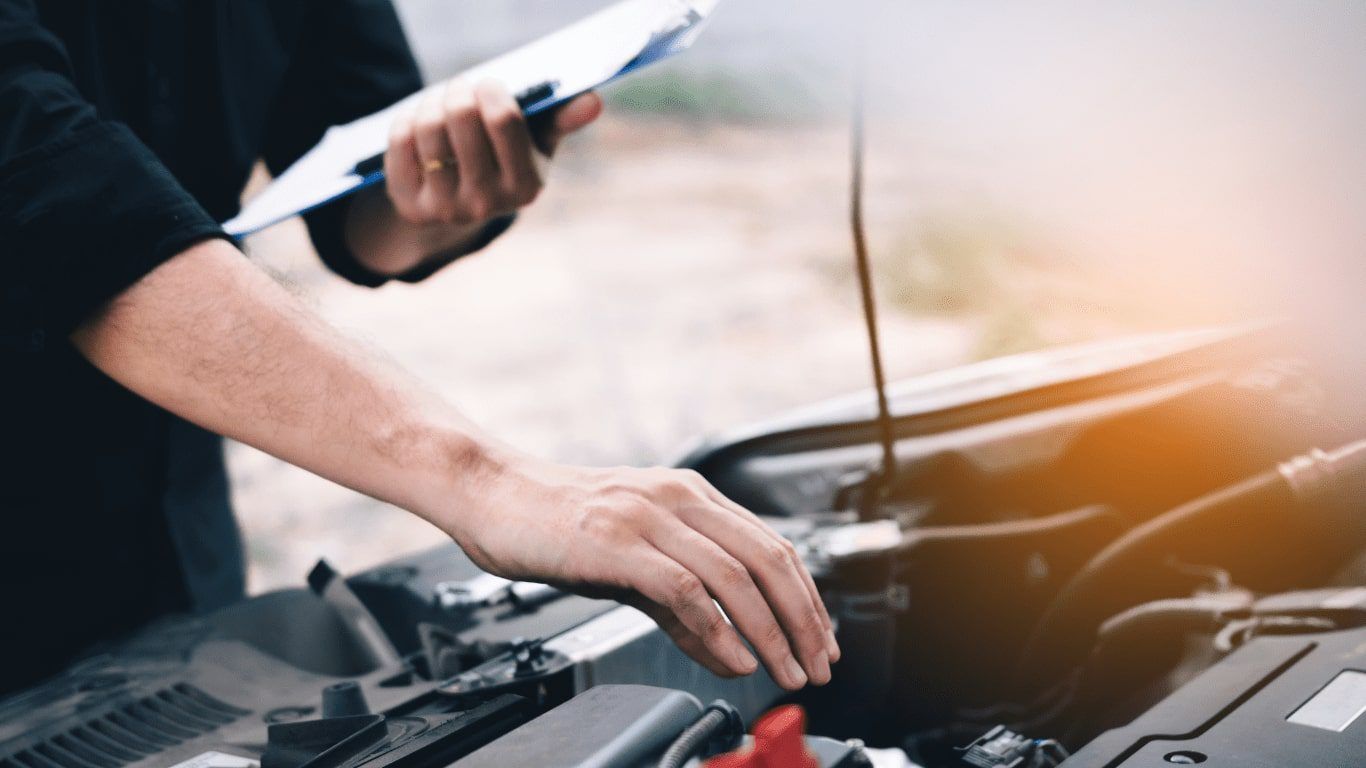 Person inspecting a car engine with a clipboard in hand, hood open outdoors