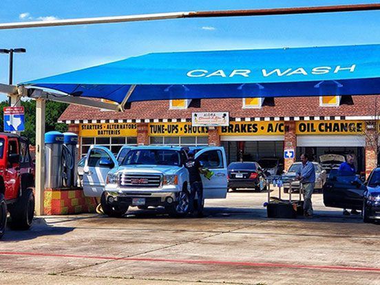 Cars lined up at a blue awning car wash and auto service station with a red wet pavement foreground