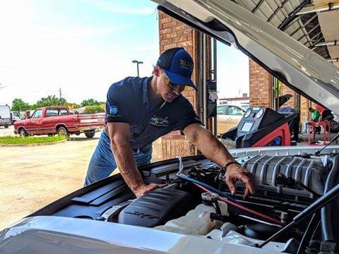 Mechanic inspecting a car engine under the hood in a garage, with a red truck outside.