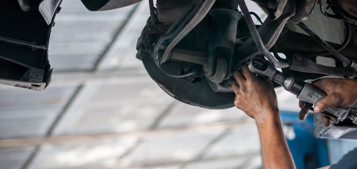 Mechanic’s hands working under a car in a garage, inspecting the underside with a tool.
