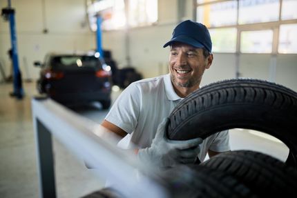 Mechanic smiling while lifting a tire in a garage, with a black car in the background