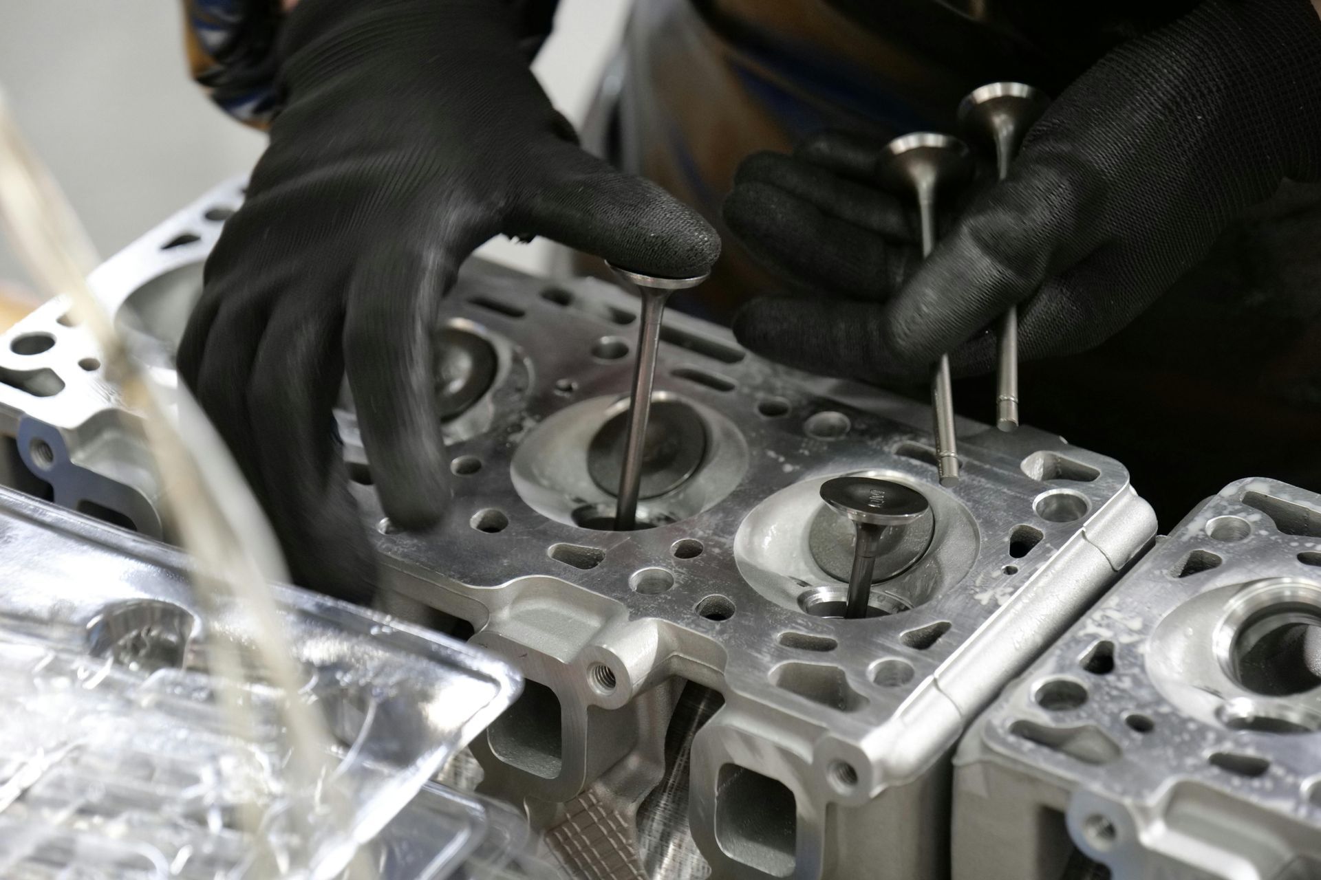 Gloved hands working on a metal engine cylinder head with tools in a workshop