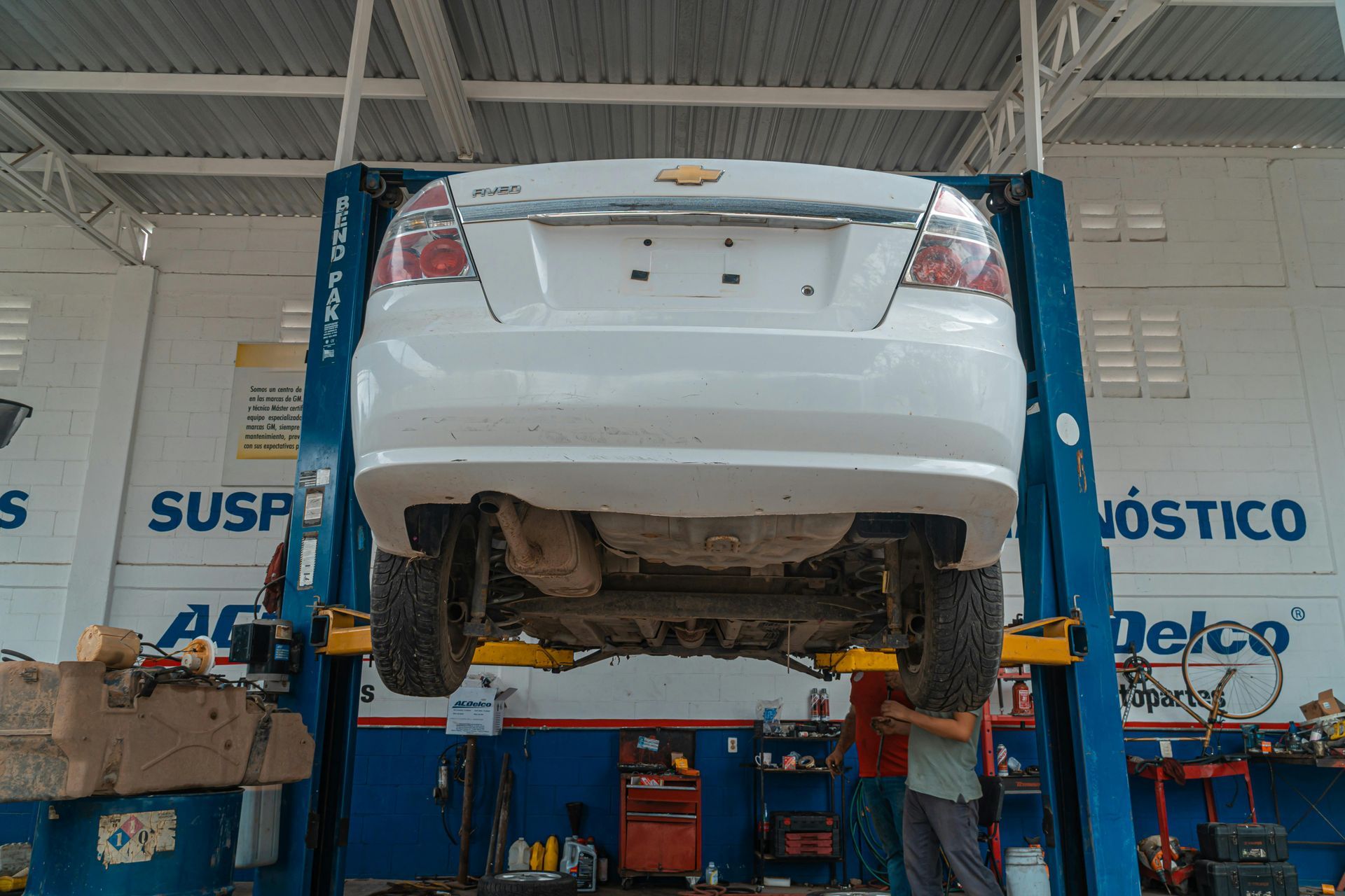White car lifted on blue hydraulic lift in auto repair shop, rear view visible under the vehicle