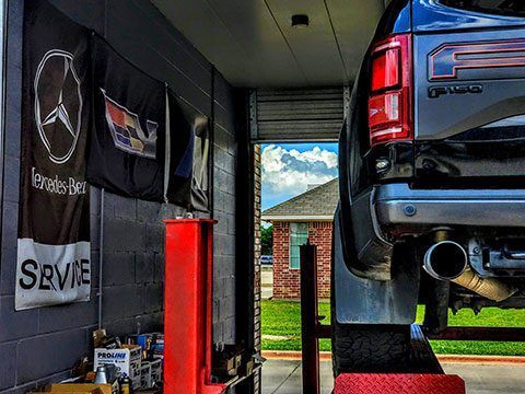 Black Mercedes pickup in a garage beside flags and a red lift, with a view of a house and yard outside.