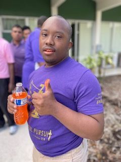 A man in a purple shirt is holding a bottle of orange soda and giving a thumbs up.