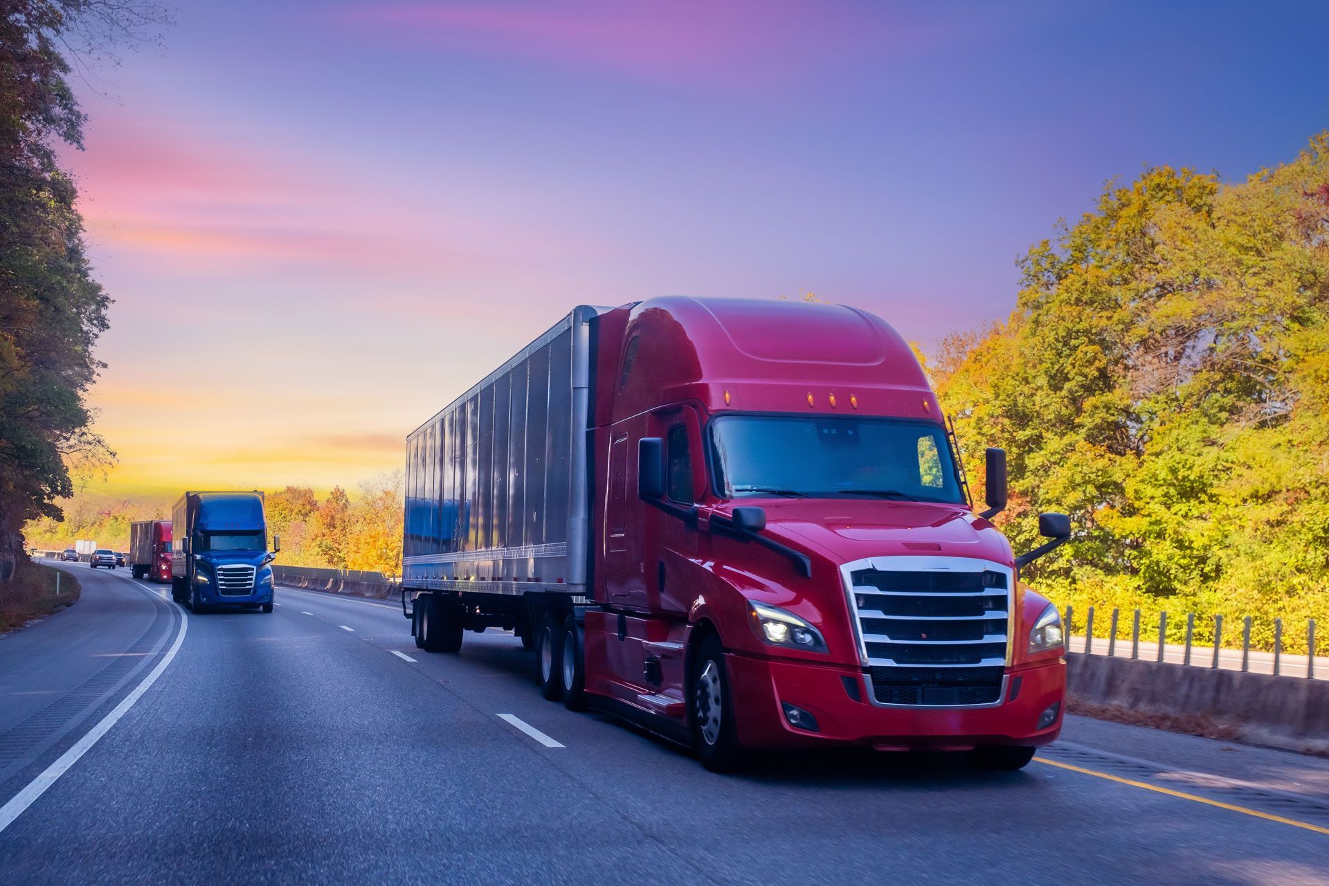 Red semi-truck driving on a highway at sunset, with another truck behind it and trees along the road.