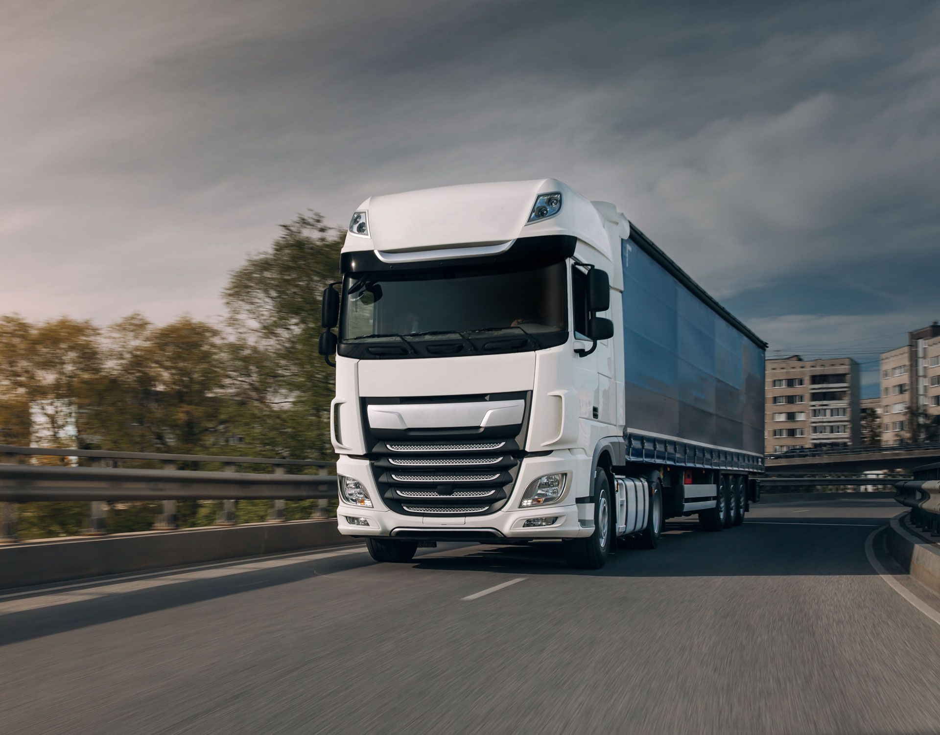 White semi-truck driving on a curved road with buildings and trees in the background