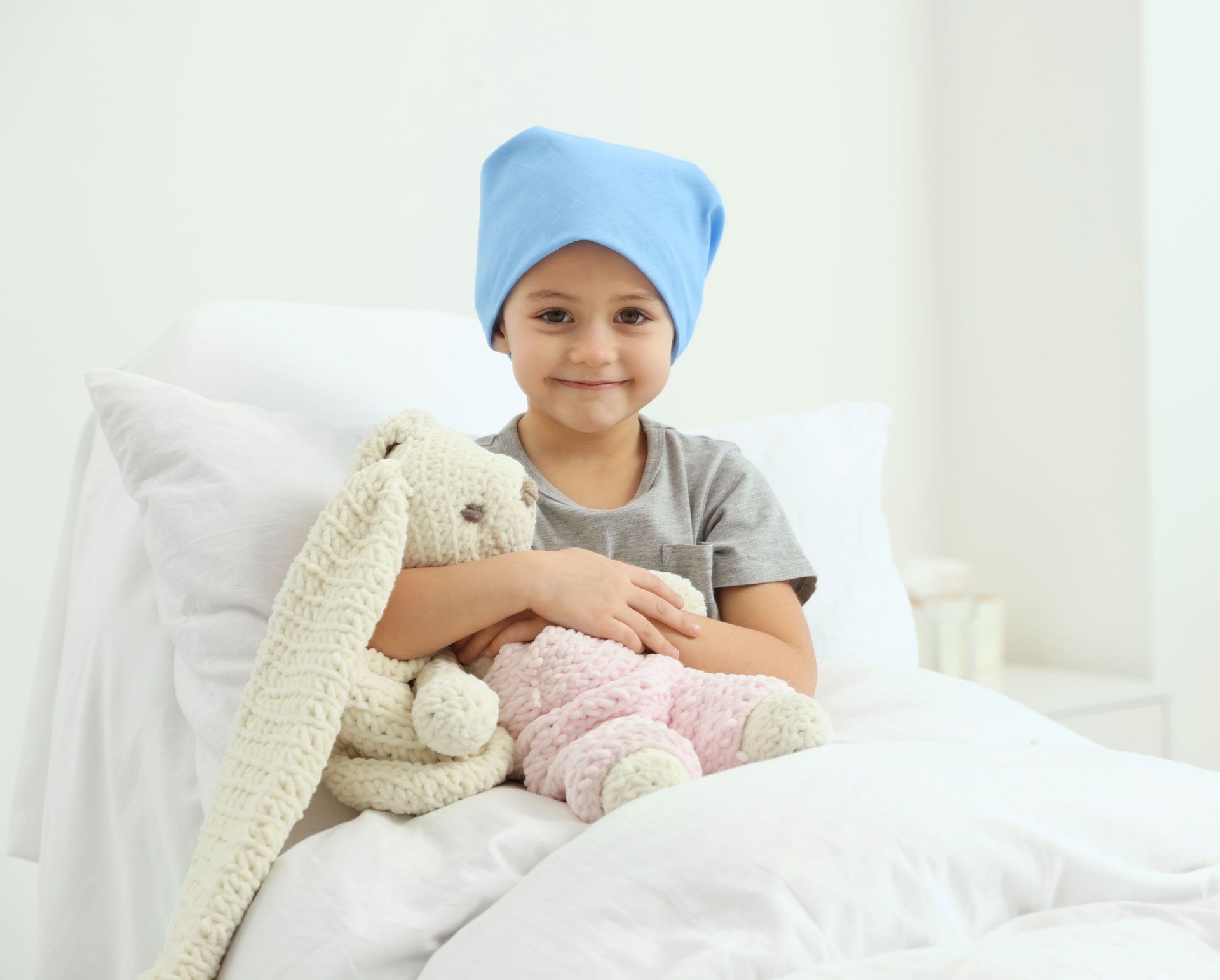 Child in a blue hat holding stuffed animals on a white bed