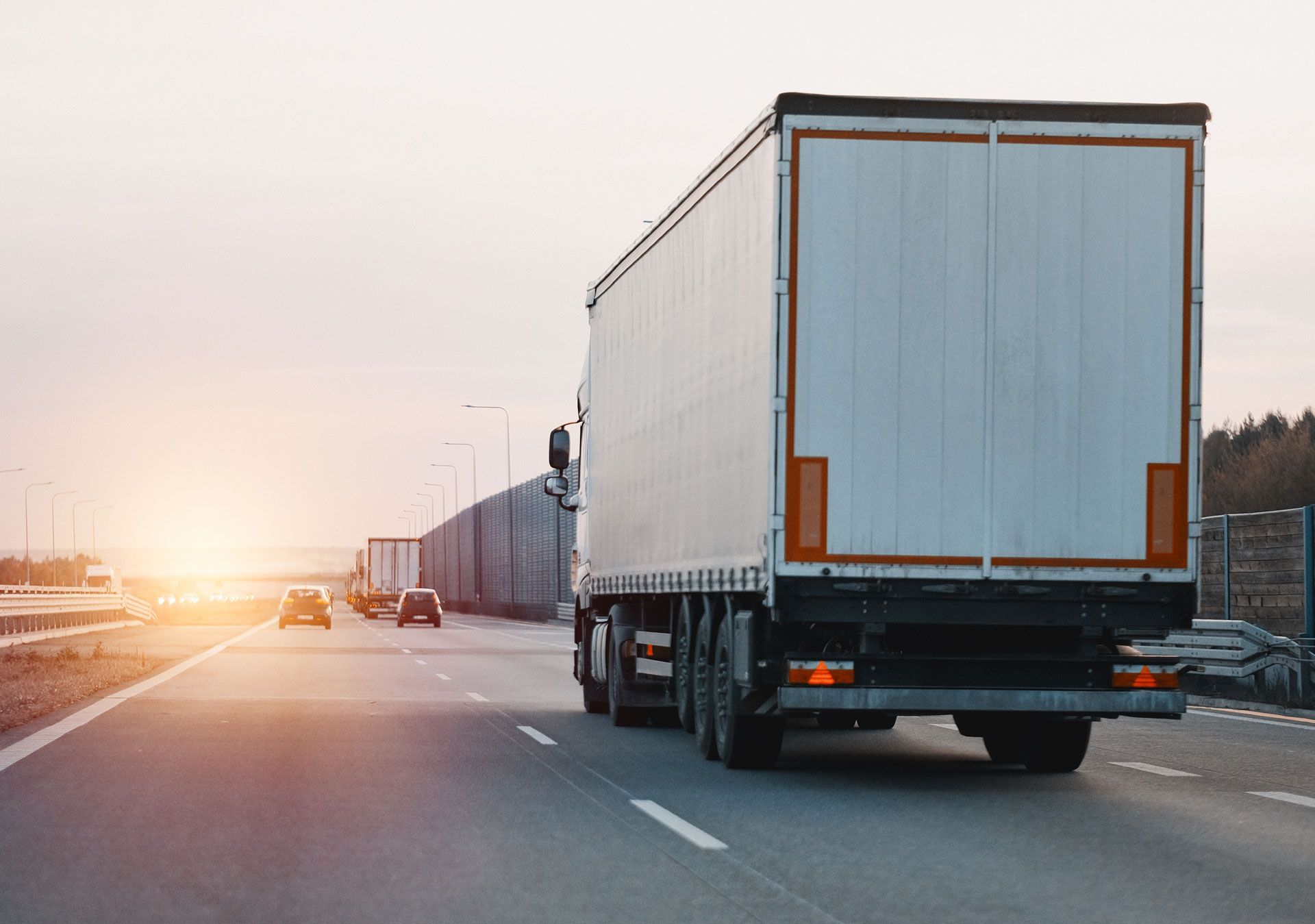 Two semi trucks driving on a highway at sunset, with a bright sky ahead.