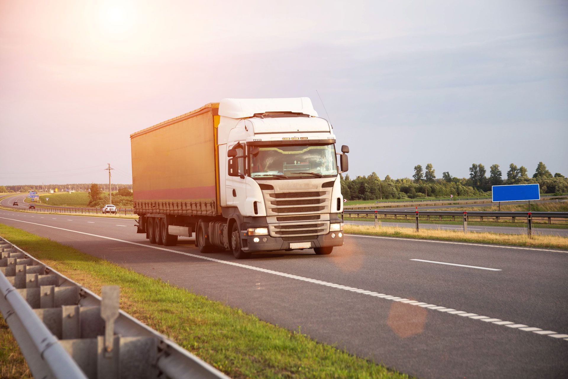 White semi-truck driving on a highway beside a grassy median and guardrail under a bright sky