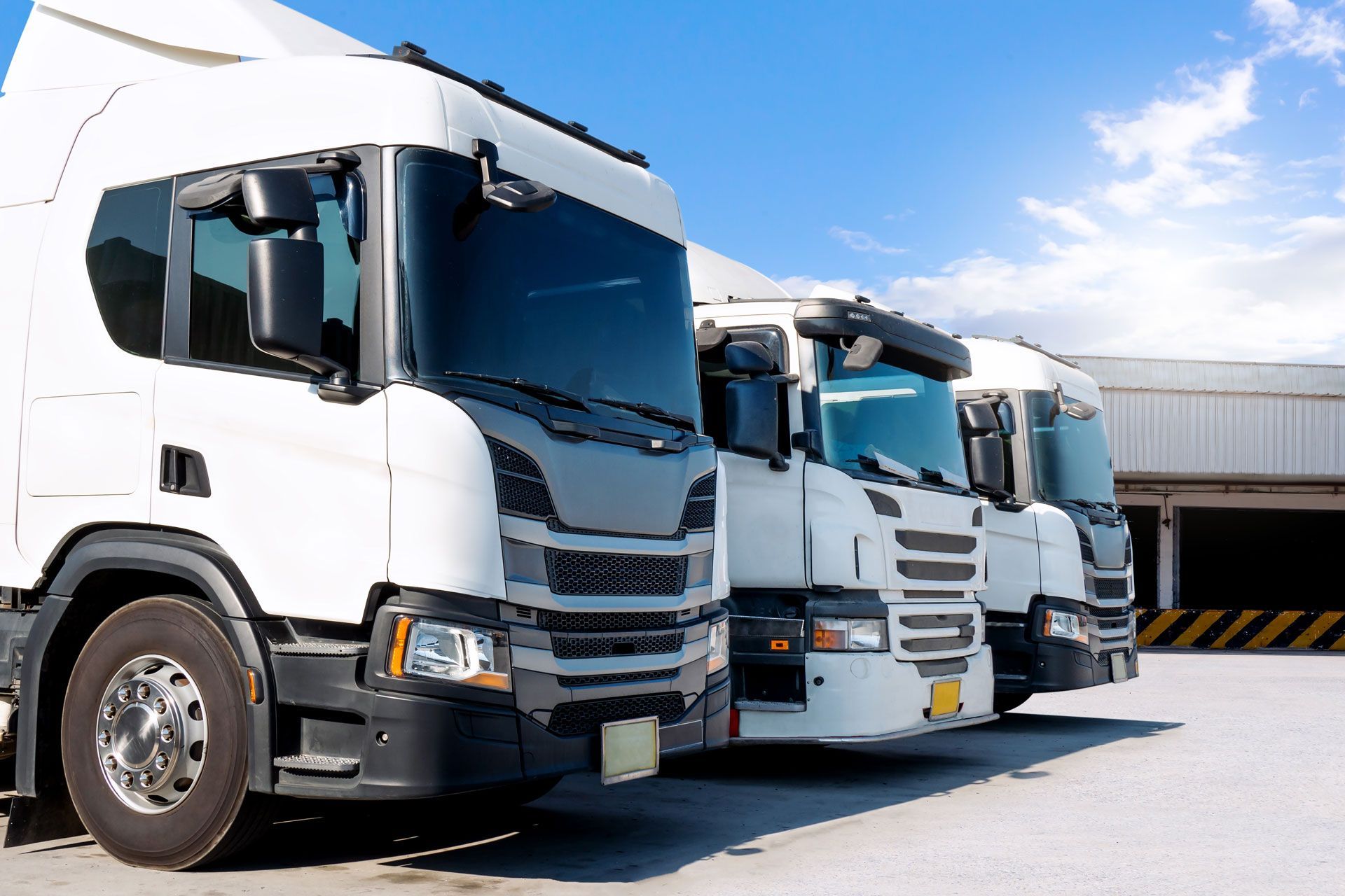 Three white semi trucks parked side by side in an industrial lot under a blue sky