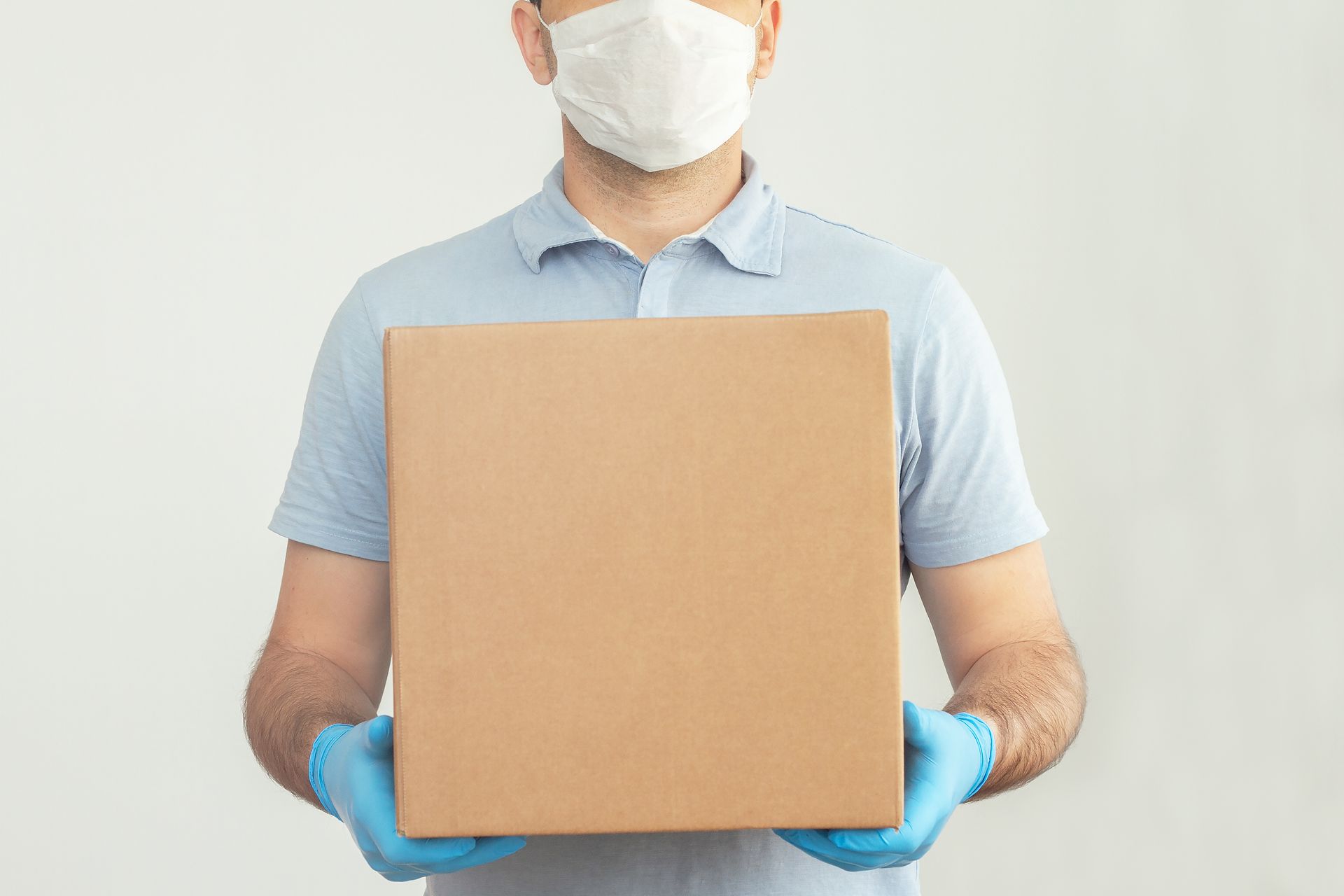 Person wearing a mask and blue gloves holding a large cardboard box against a white background