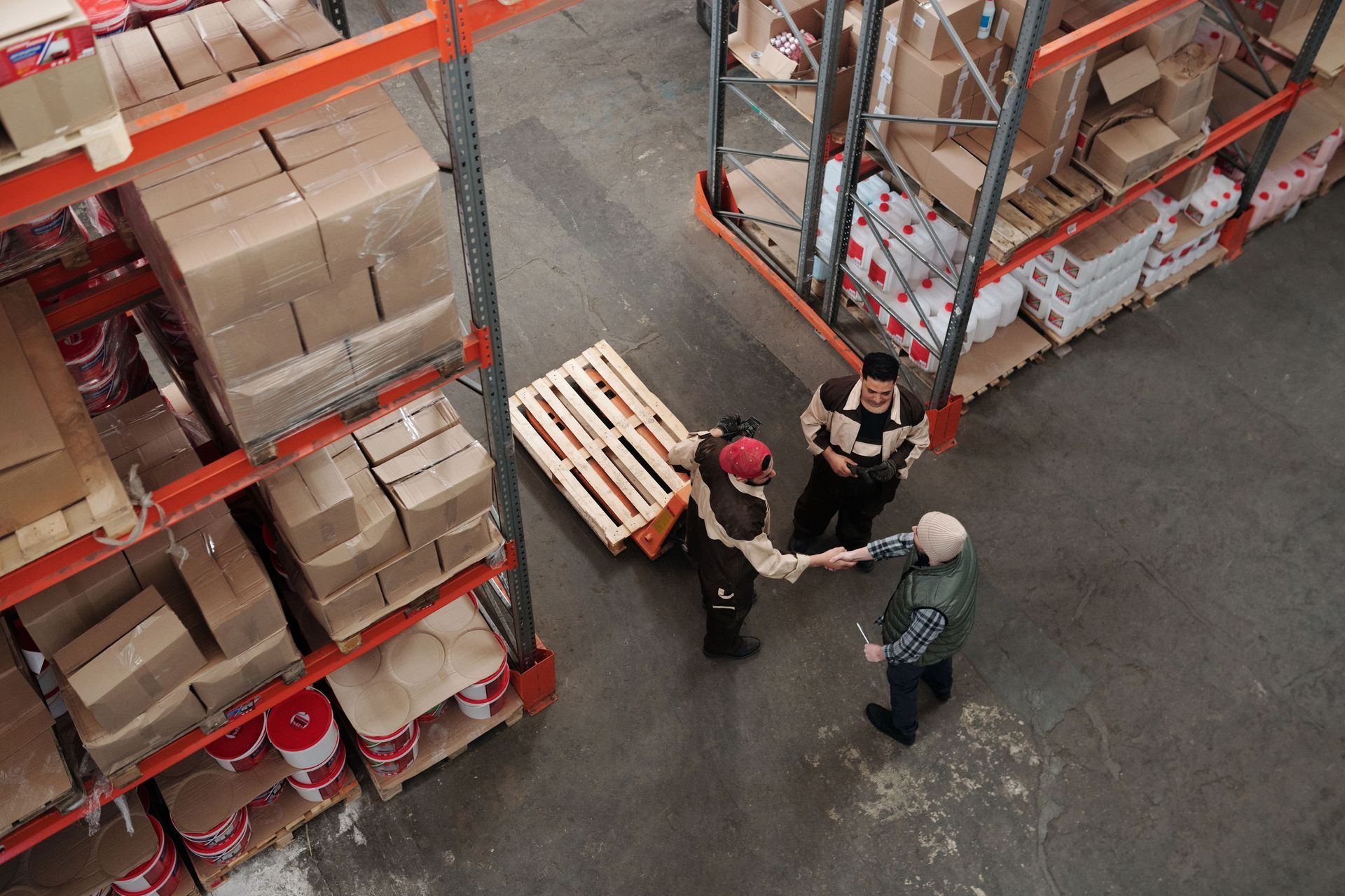 Four workers in a warehouse stand in a small circle near stacked pallets and shelves.